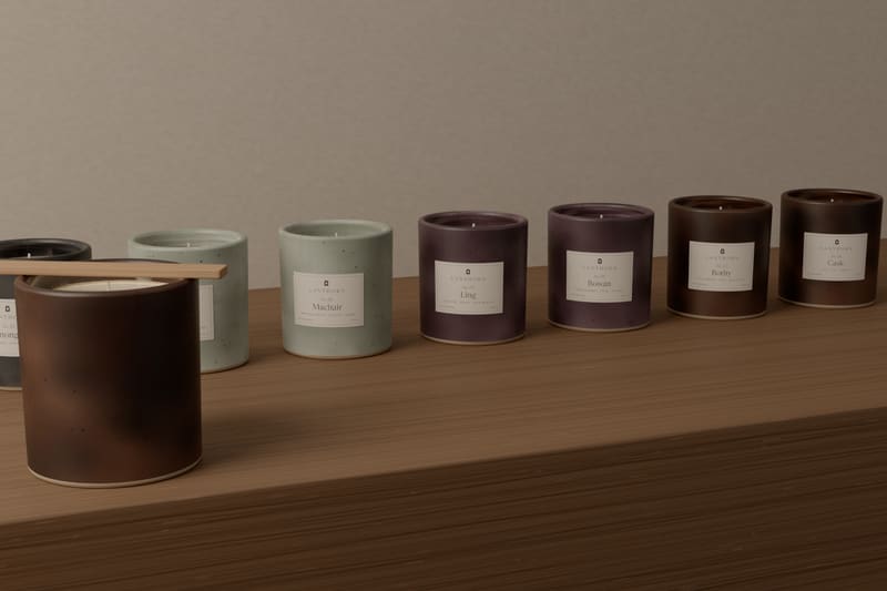 A long oak bench in the workshop with eight candles in a row, one of each scent, their labels facing out, beside a freshly poured candle held straight by a wooden bar.