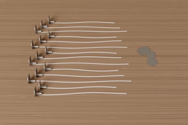 Fourteen flat-braided cotton wicks with round steel sustainer tabs, laid out in parallel on an oak board, with a few spare tabs beside them.