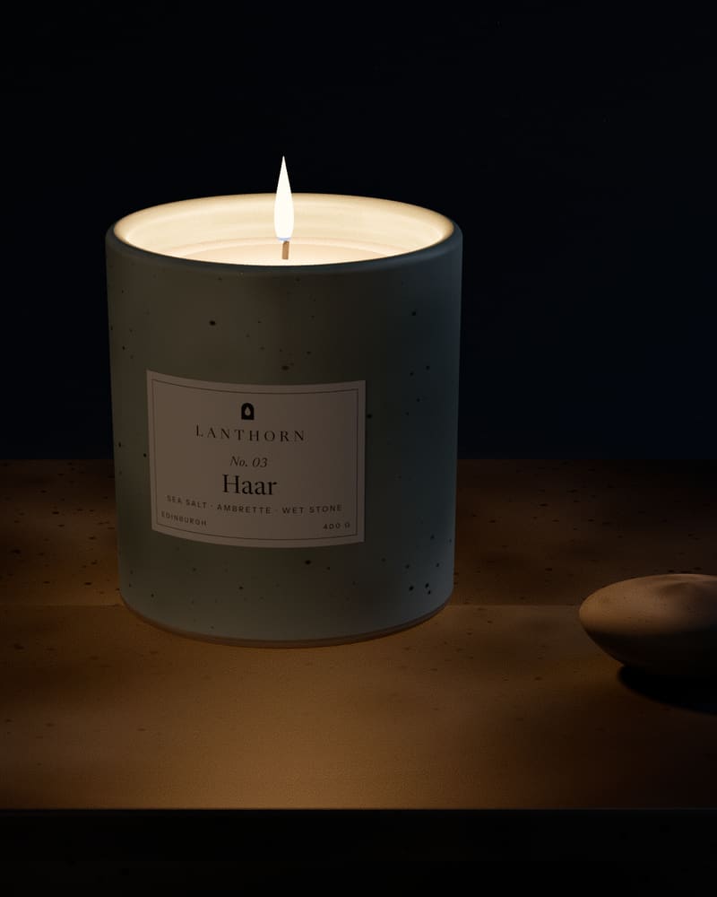 Haar candle lit in its pale grey-green stoneware vessel, set on a slab of grey slate with a smooth beach pebble.
