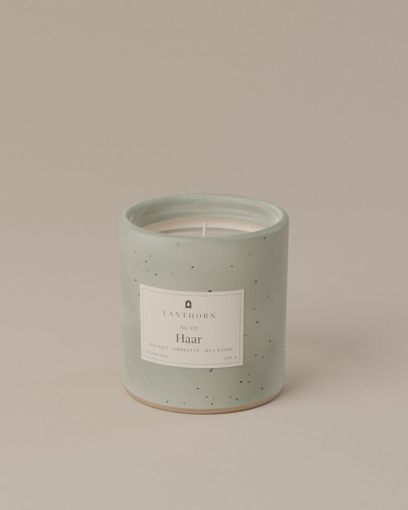 Haar 220 g candle in pale grey-green speckled stoneware with its paper label, on a plain warm-grey backdrop.